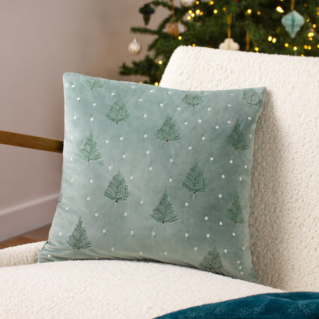 Square velvet cushion in mint green with embroidered fir trees and scattered pale dots, shown on a cream sherpa armchair beside a lit Christmas tree.