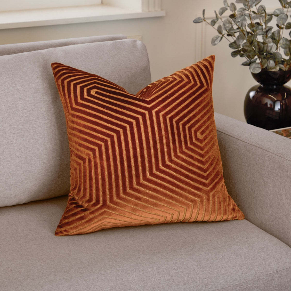 Evoke Geometric Velvet Cushion in Brick, a square cut-velvet cushion with radiating angular channel patterns in rust and burnt orange, shown on a light grey sofa.