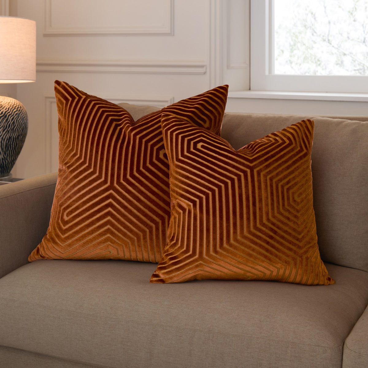Two Evoke Geometric Velvet Cushions in rust and burnt orange with radiating cut-velvet channel patterns, styled on a beige sofa in a living room.