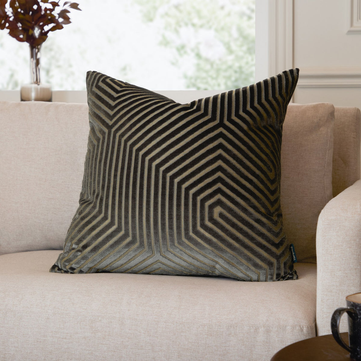 Evoke Geometric Velvet Cushion in charcoal with gold-toned hexagonal pattern, shown on a beige sofa in a living room setting.