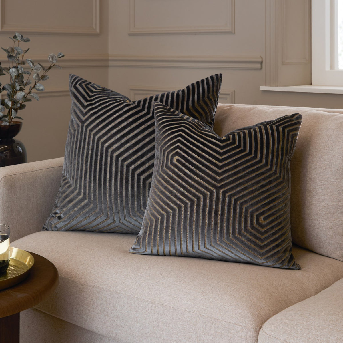 Two Evoke Geometric Velvet Cushions in charcoal with cut velvet hexagonal line patterns, styled on a beige sofa in a living room setting.