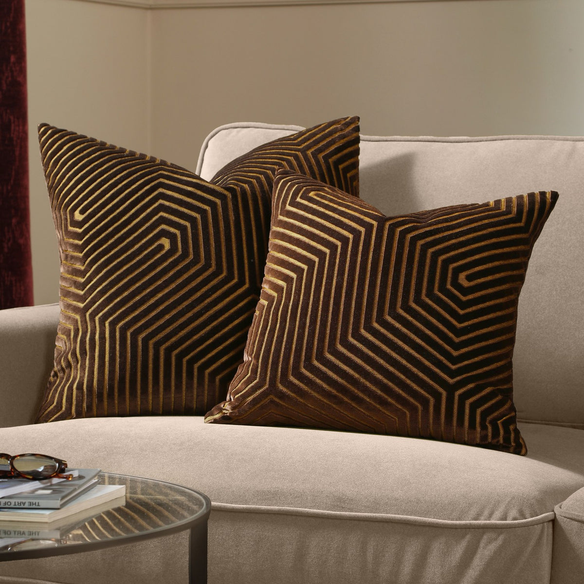 Two Evoke Geometric Velvet Cushions in chocolate brown with tan and gold chevron patterning, styled on a beige sofa beside a glass side table.