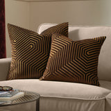 Two Evoke Geometric Velvet Cushions in chocolate brown with tan and gold chevron patterning, styled on a beige sofa beside a glass side table.