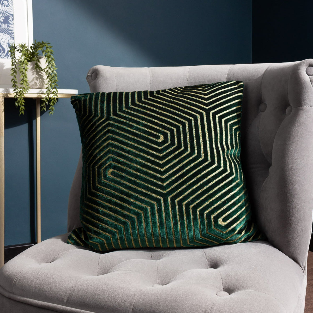 Evoke Geometric Velvet Cushion in emerald with gold chevron pattern, shown on a grey tufted armchair in a living room setting.