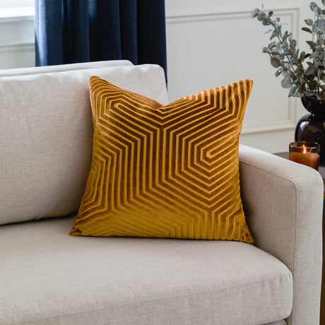 Evoke Geometric Velvet Cushion in gold with a cut-pile chevron pattern, shown on a beige sofa in a living room setting.