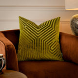 Evoke square velvet cushion in olive and chartreuse with a geometric chevron pile pattern, shown on a rust velvet armchair beside a table lamp.