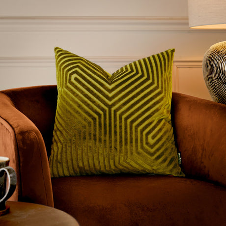 Evoke square velvet cushion in olive and chartreuse with a geometric chevron pile pattern, shown on a rust velvet armchair beside a table lamp.