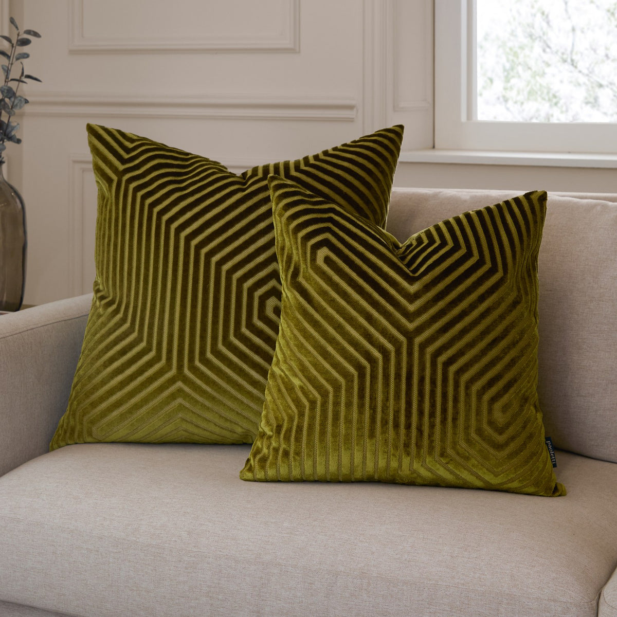 Two Evoke square velvet cushions in olive and chartreuse with a geometric chevron pattern, shown on a beige sofa in a living room setting.