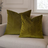 Two Evoke square velvet cushions in olive and chartreuse with a geometric chevron pattern, shown on a beige sofa in a living room setting.