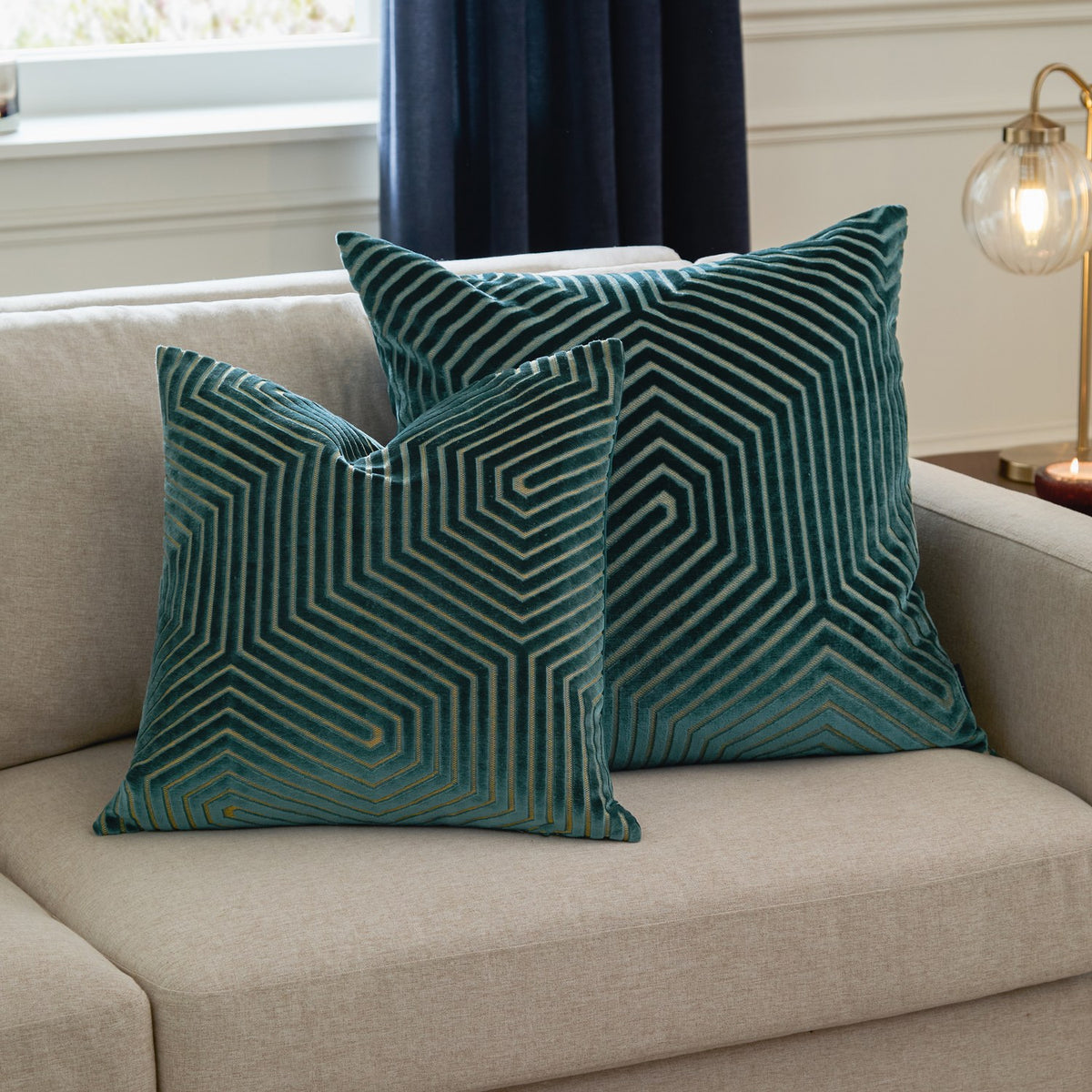 Two Evoke Geometric Velvet Cushions in teal with cut-pile chevron and hexagon patterns and metallic gold thread accents, styled on a beige sofa.