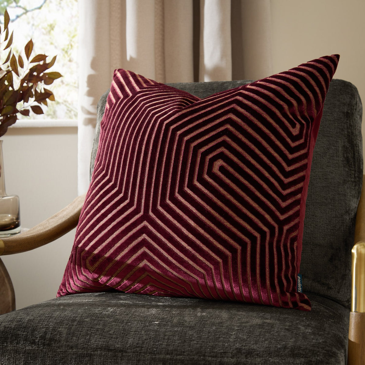 Evoke Geometric Velvet Cushion in burgundy with gold-toned chevron stitching, shown square-shaped on a dark grey armchair in a living room setting.
