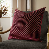 Evoke Geometric Velvet Cushion in burgundy with gold-toned chevron stitching, shown square-shaped on a dark grey armchair in a living room setting.