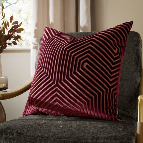 Evoke Geometric Velvet Cushion in burgundy with gold-toned chevron stitching, shown square-shaped on a dark grey armchair in a living room setting.