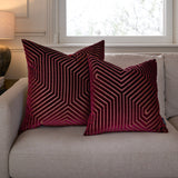 Two square Evoke velvet cushions in burgundy with raised geometric chevron patterning, styled on a beige sofa beside a window.