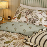 Fable Reversible Animal Duvet Cover Set in Willow Green on a bed with cream woodland-print pillowcases featuring foxes, hares and trees, plus a sage leaf-sprig bolster and striped cushions.