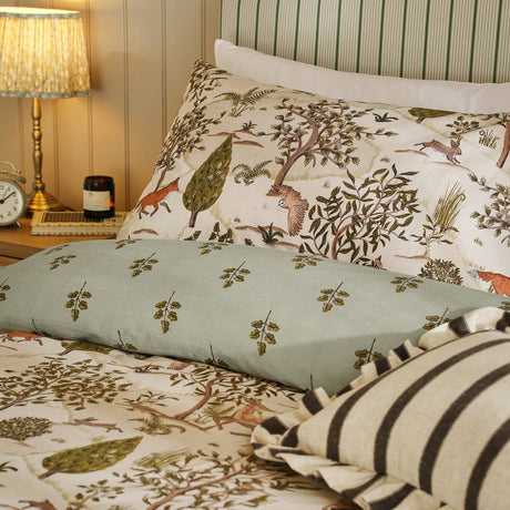 Fable Reversible Animal Duvet Cover Set in Willow Green on a bed with cream woodland-print pillowcases featuring foxes, hares and trees, plus a sage leaf-sprig bolster and striped cushions.
