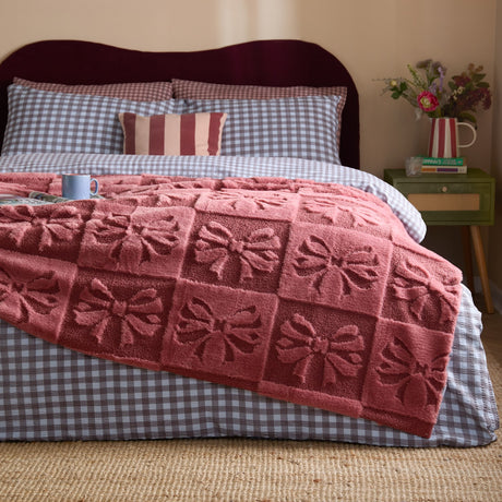 Pink faux fur throw with raised bow pattern draped over a bed with grey gingham bedding, styled in a bedroom setting.