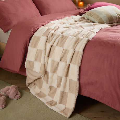 Fur Boucle Check Velvet Faux Fur Throw in natural draped on a pink bed, showing a beige and cream checkerboard pattern in mixed boucle and plush textures.