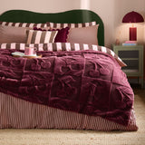Morello faux fur throw in deep purple on a bed, showing a boucle checkerboard texture with a raised cherry pattern, styled with striped bedding in a bedroom setting.