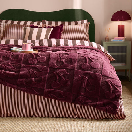 Morello faux fur throw in deep purple on a bed, showing a boucle checkerboard texture with a raised cherry pattern, styled with striped bedding in a bedroom setting.
