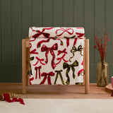 Nathaliefestivebows polyester throw draped over a wooden stand, cream fabric printed with berry red and pine green bows, some in tartan check, styled with dried flowers in a glass vase.