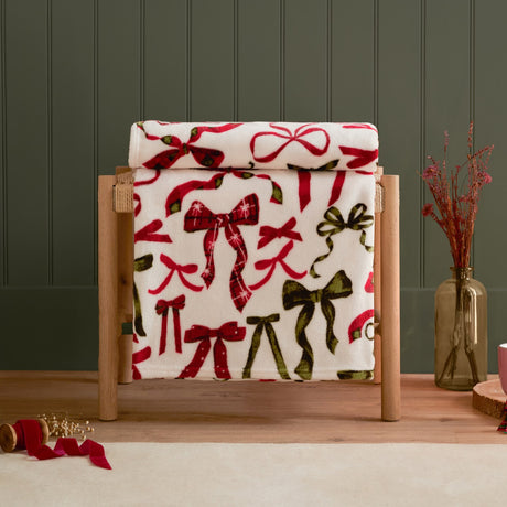 Nathaliefestivebows polyester throw draped over a wooden stand, cream fabric printed with berry red and pine green bows, some in tartan check, styled with dried flowers in a glass vase.
