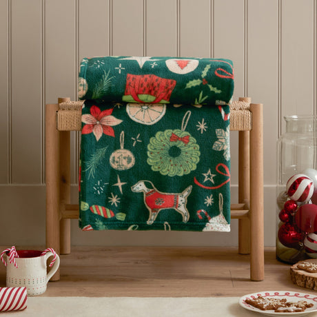 Festive Treats Printed Polyester Throw in emerald green, draped over a wooden rack with a Christmas print of dogs, wreaths, ornaments and candy canes, styled in a living room setting.