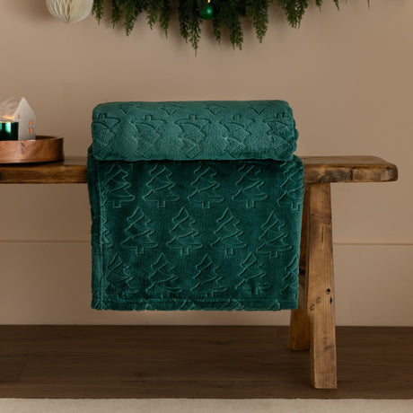 Festive Trees Woodland Polyester Throw in emerald green, folded over a wooden bench, showing an embossed Christmas tree pattern raised from the fleece pile.