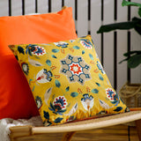 Folk Flora Floral Polyester Cushion in ochre-yellow with a folk-art medallion, floral and leaf print in teal, red and cream, shown propped on an orange sofa.