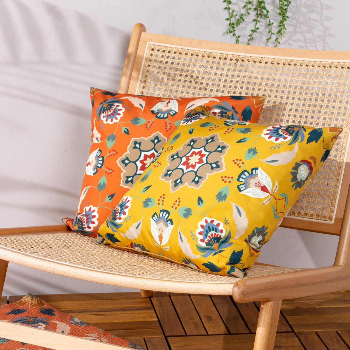 Folk Flora Floral Polyester Cushion in ochre-yellow with a folk-art medallion print, styled on a rattan-backed wooden chair alongside an orange floral cushion.
