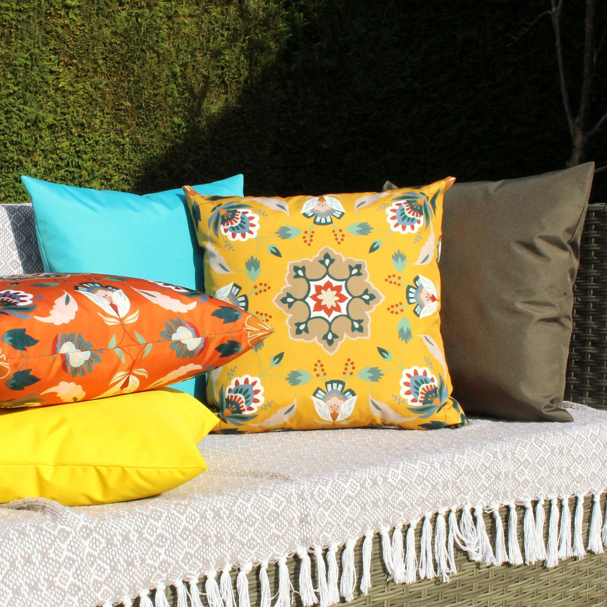 Folk Flora Floral Polyester Cushion in ochre-yellow with a folk-art medallion and floral pattern, shown among assorted cushions on an outdoor sofa.