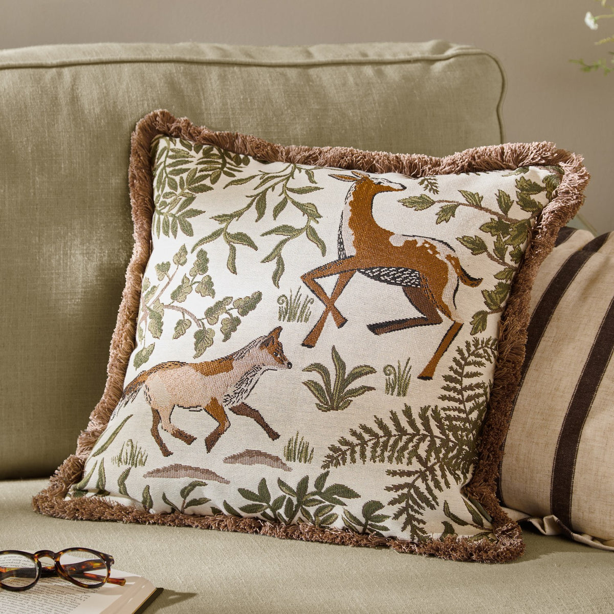 Fable Fox and Deer Woodland Cotton Cushion in Willow Green, square with dusky-pink fringe trim, shown on a sage sofa with cream and brown striped cushions.