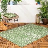 Forage Garden Floral Polyester Rug in sage, shown on an outdoor wooden deck with a paneled cream floral, fern and peacock pattern amid potted plants.