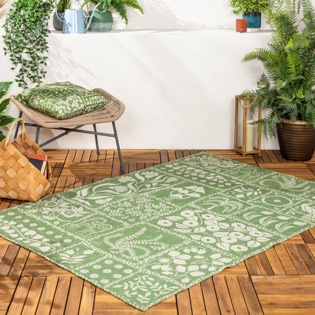 Forage Garden Floral Polyester Rug in sage, shown on an outdoor wooden deck with a paneled cream floral, fern and peacock pattern amid potted plants.