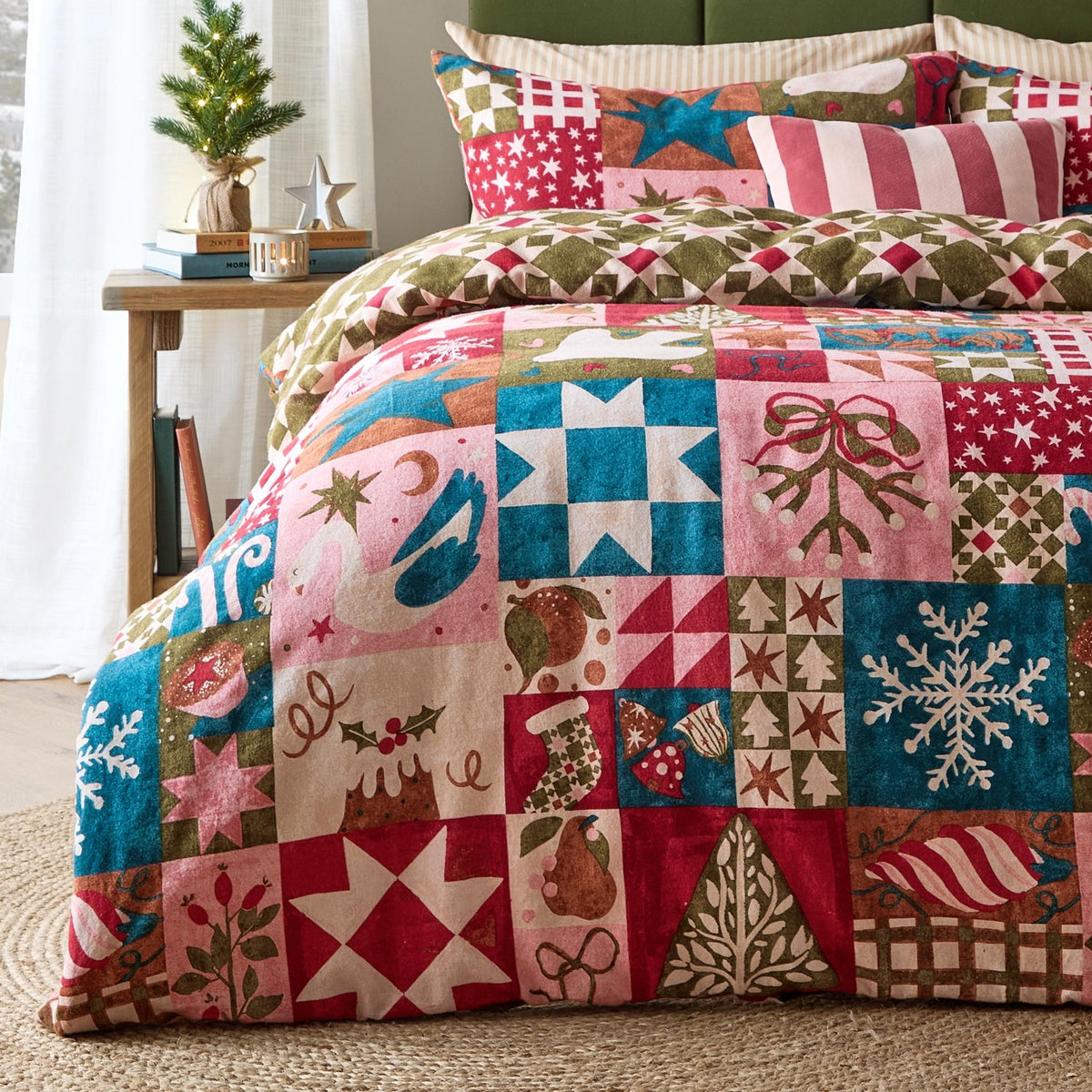 Fika Cotton Geometric Duvet Cover Set in Multi dressing a bed, featuring a reversible Christmas patchwork of stars, snowflakes and swans in pink, teal, red and olive, styled in a bedroom.