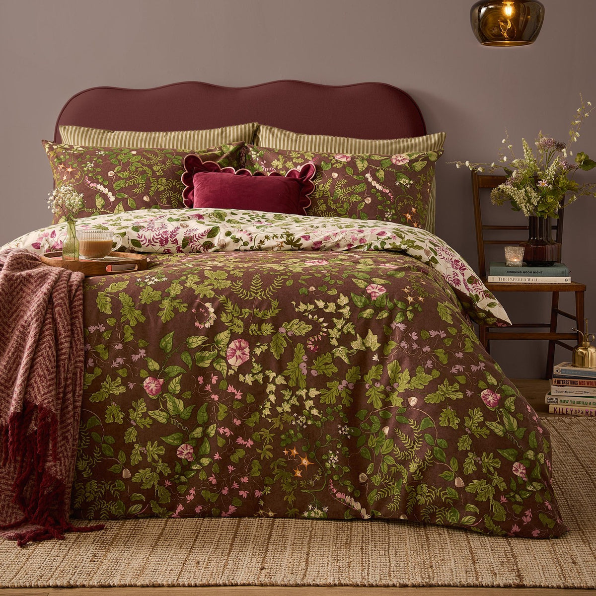 Flora Wilde Reversible Floral Duvet Cover Set in Chocolate on a bed with pink and green wildflower print, styled with striped pillowcases and a burgundy velvet cushion in a bedroom.