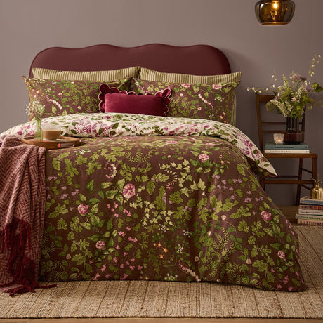Flora Wilde Reversible Floral Duvet Cover Set in Chocolate on a bed with pink and green wildflower print, styled with striped pillowcases and a burgundy velvet cushion in a bedroom.