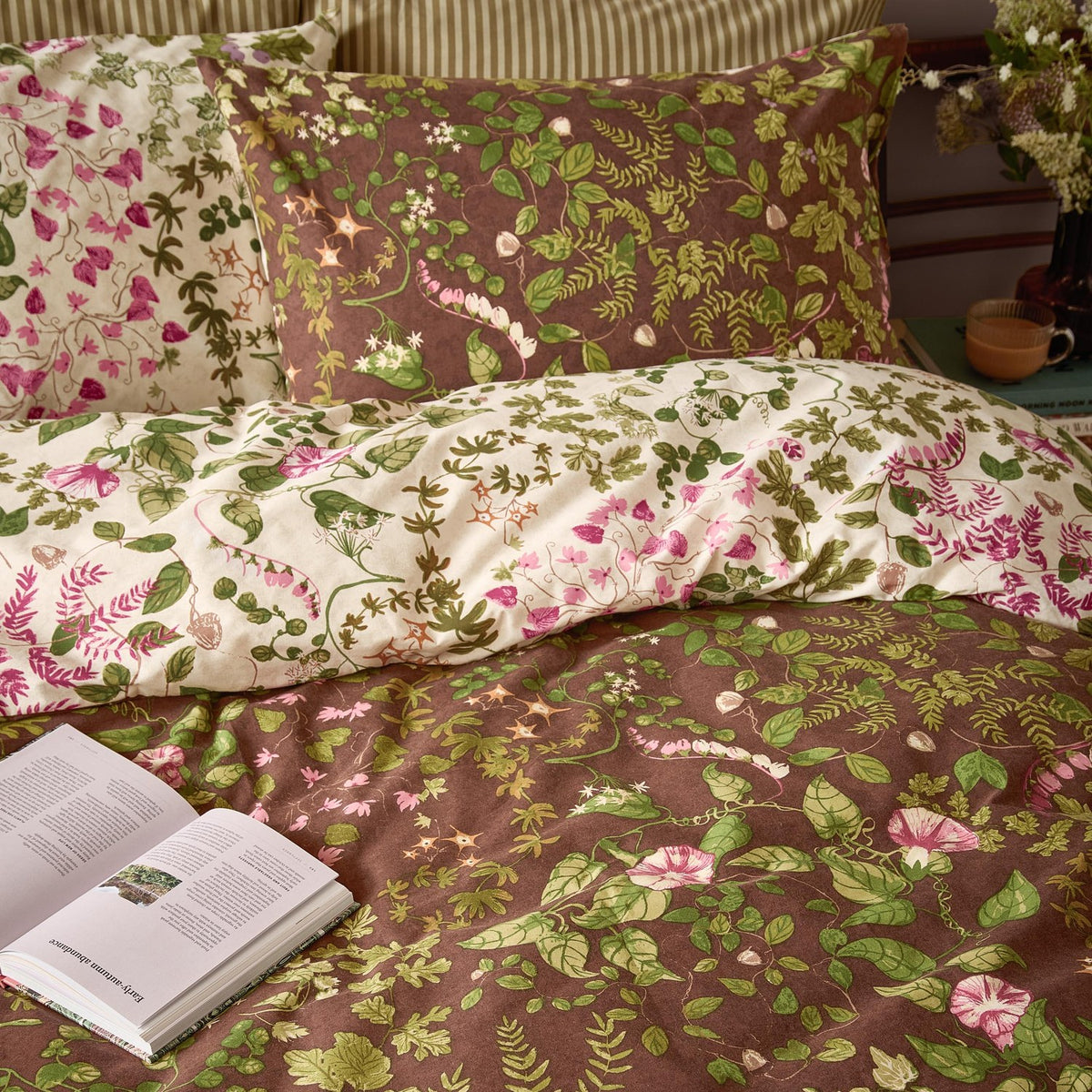 Flora Wilde reversible duvet cover set on a bed, showing floral vine and leaf print in pink and green on chocolate brown, paired with matching pillowcases in cream and brown, styled with a book.