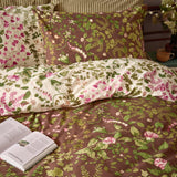 Flora Wilde reversible duvet cover set on a bed, showing floral vine and leaf print in pink and green on chocolate brown, paired with matching pillowcases in cream and brown, styled with a book.