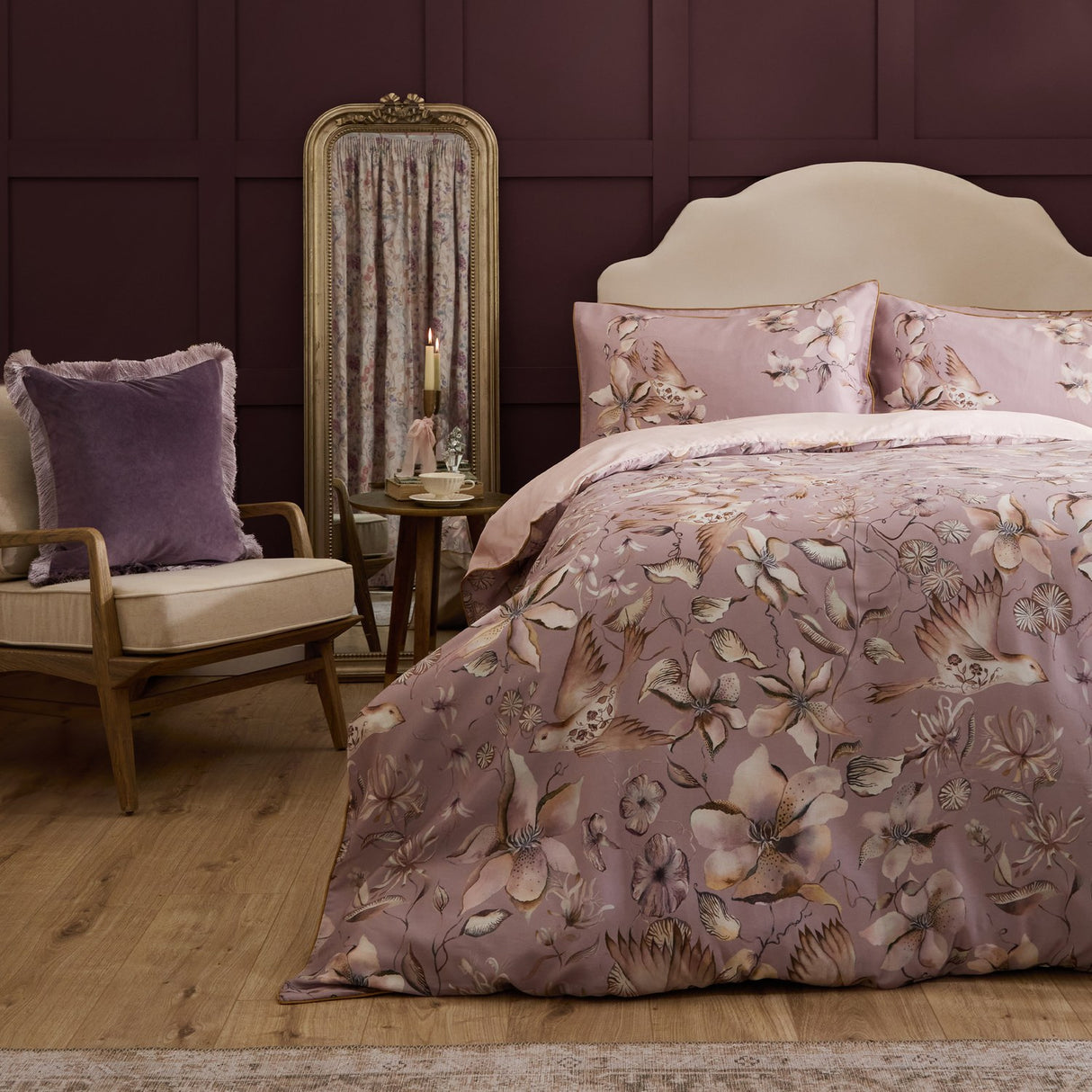 Floella Cotton Botanical Duvet Cover Set in Purple, dressed on a bed with floral and bird print pillowcases, styled in a bedroom with a velvet armchair and gilt mirror.