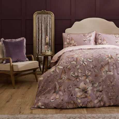 Floella Cotton Botanical Duvet Cover Set in Purple, dressed on a bed with floral and bird print pillowcases, styled in a bedroom with a velvet armchair and gilt mirror.