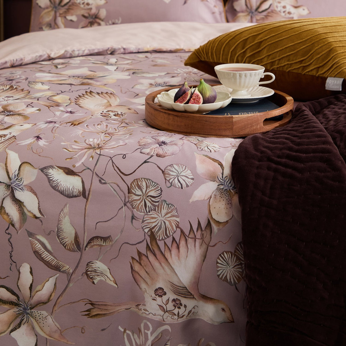Floella Cotton Botanical Duvet Cover Set in purple, styled on a bed with a floral bird print in cream and mauve, mustard cushion and dark throw with a breakfast tray.