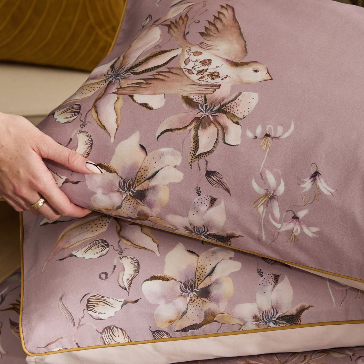 Floella Cotton Botanical Duvet Cover Set in purple, showing pillowcases with watercolour-style bird and floral print, edged in mustard piping, styled with a hand smoothing the fabric.