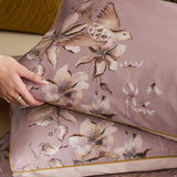 Floella Cotton Botanical Duvet Cover Set in purple, showing pillowcases with watercolour-style bird and floral print, edged in mustard piping, styled with a hand smoothing the fabric.