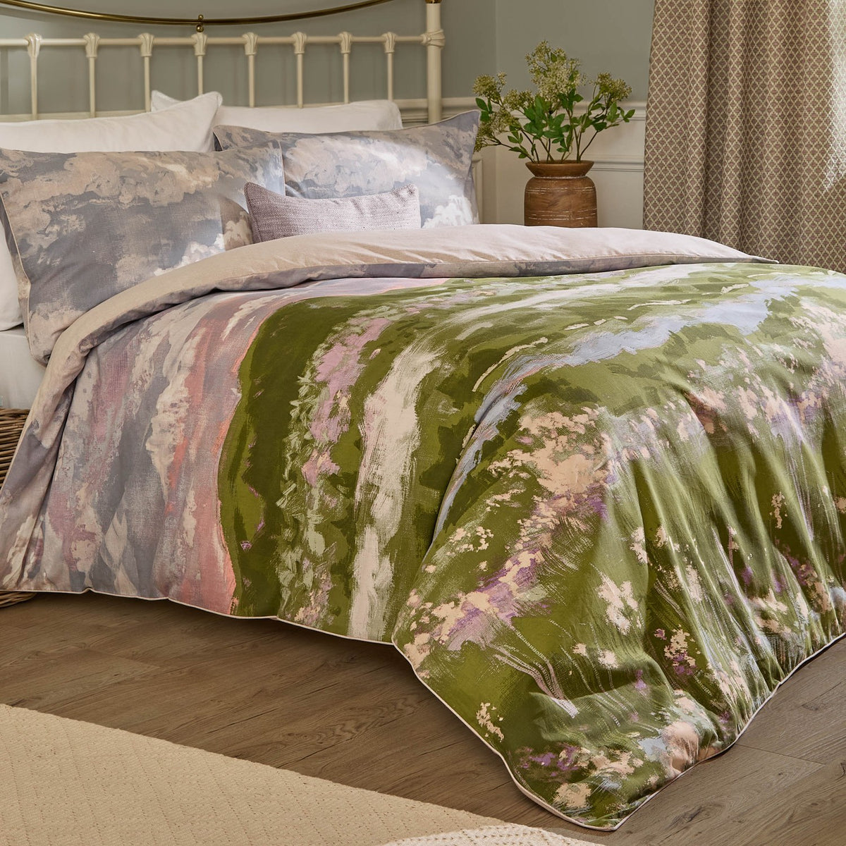 Foinaven Cotton Piped Duvet Cover Set in Heather with a painterly landscape print in mossy green, dusky pink and neutral tones, paired with cloud-print pillowcases on a bed.