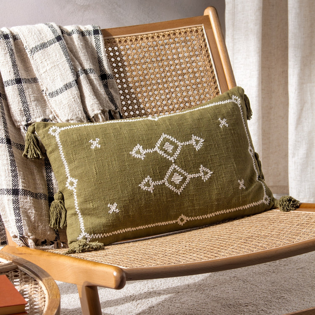 Folis Geometric Cotton Cushion in khaki with cream embroidered diamond motifs and corner tassels, styled on a rattan chair with a checked throw.