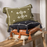 Folis Geometric Cotton Cushion in khaki green with cream embroidered diamond motifs and side tassels, stacked with navy, orange and cream cushions on a wooden bench.