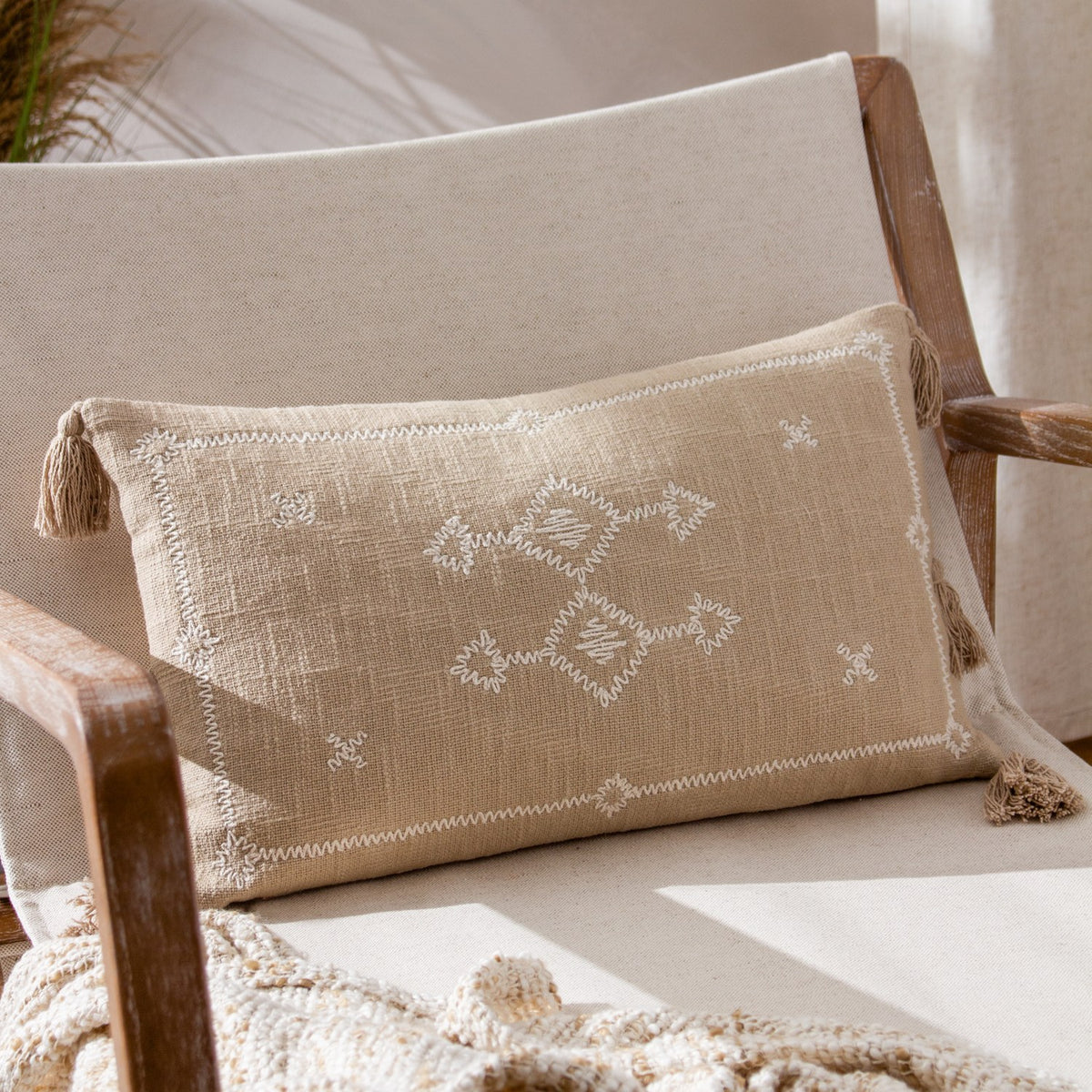 Folis Geometric Cotton Cushion in stone, a rectangular lumbar cushion with cream diamond and cross embroidery and corner tassels, styled on a wooden armchair.