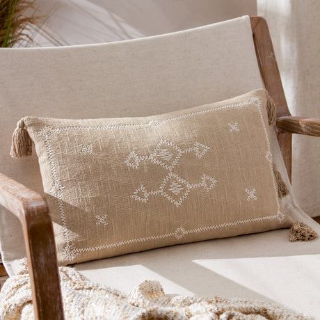 Folis Geometric Cotton Cushion in stone, a rectangular lumbar cushion with cream diamond and cross embroidery and corner tassels, styled on a wooden armchair.