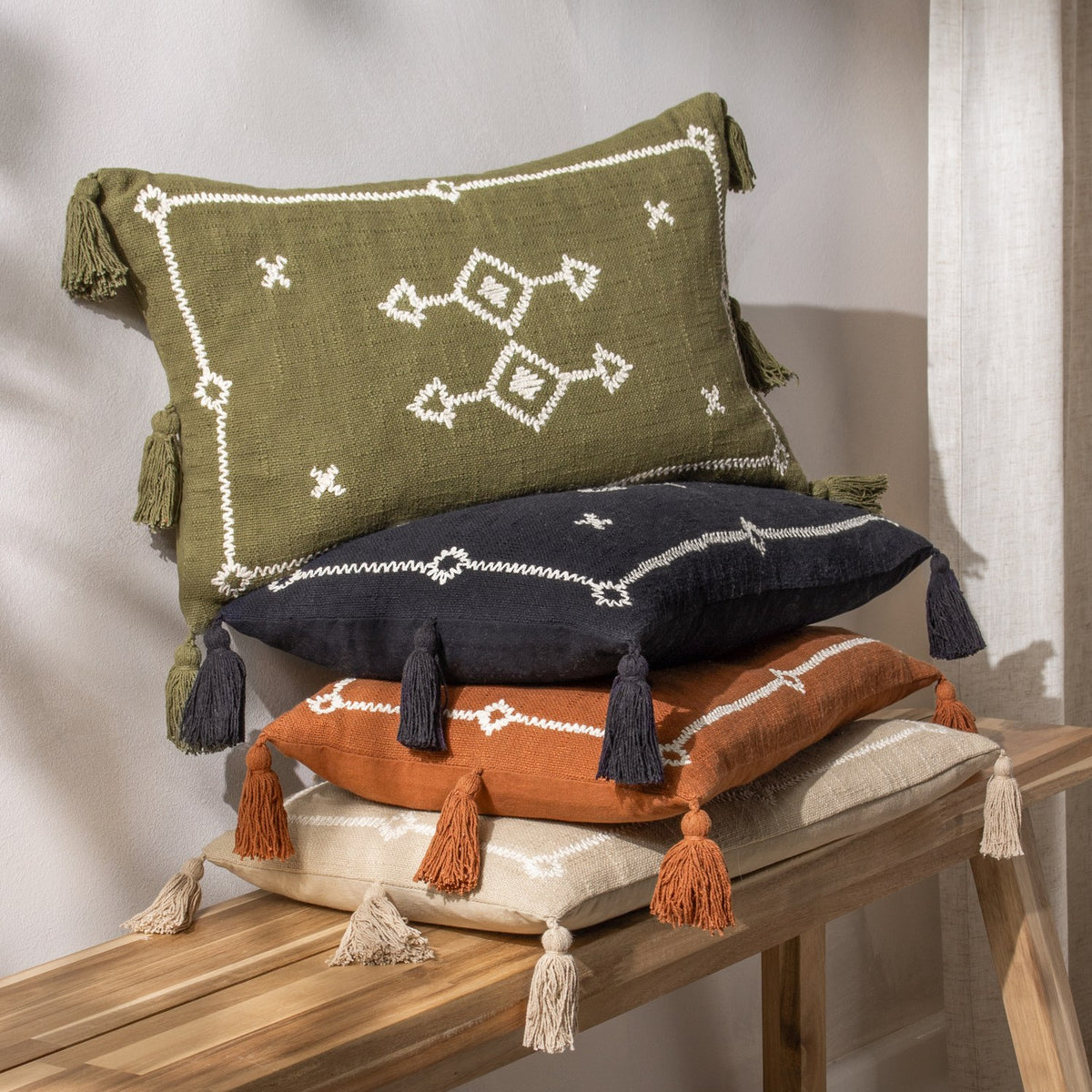 Folis Geometric Cotton Cushion in stone with cream embroidered diamond motif and corner tassels, shown stacked with black, orange and cream cushions on a wooden bench.